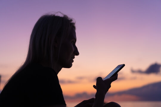 Happy young woman with a mobile phone on the background of a beautiful rose-lilac sunset on the sea. Concept of modern technologies