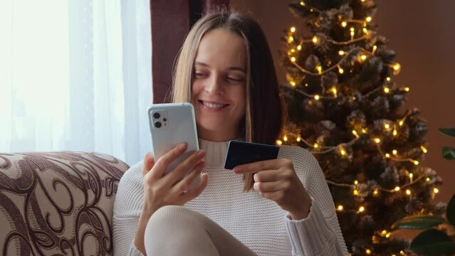 Christmas shopping. Celebration New year. Cheerful brown haired woman entering data from credit card in cellphone for paying for gifts sitting on sofa near X-mas tree.