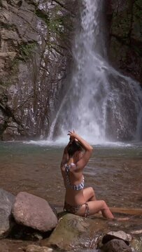 Beautiful young filipino woman in bikini sitting next to a waterfall fixing her hair. Vertical video format.