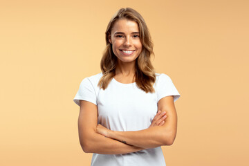 Attractive woman wearing white t shirt, arms crossed standing isolated on beige background