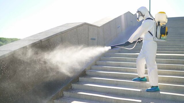 A male disinfectant in a white protective suit disinfects city streets, kills viruses with equipment. Epidemic, coronavirus, covid.