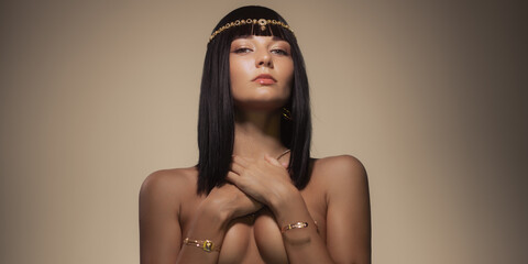 Beauty fashion portrait of naked woman queen cleopatra style covering her breasts by hands, looking at camera, posing bare with perfect make-up and golden jewelry  - obrazy, fototapety, plakaty