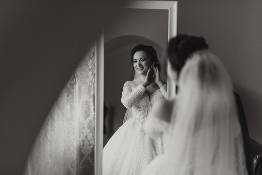 The bride in a wedding dress poses in her room, looking in the mirror. Black and white photo. Morning of the bride. Preparation. Wedding photography