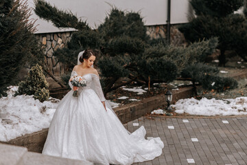 Fototapeta premium The bride in a white dress with a train and a long veil poses in nature, holding a bouquet. Walk. Winter wedding in nature.