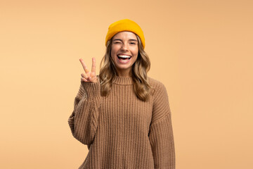 Portrait of smiling beautiful woman wearing stylish yellow hat and winter brown sweater showing victory sign looking at camera isolated on beige background, copy space. Concept of advertisement