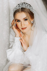 A bride in a silk suit with a veil poses in her room, sitting on the floor next to her wedding dress. Morning of the bride. Preparation