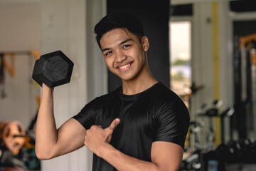A young handsome asian man doing standing single arm bicep curls while pointing to himself. Working out arms at the gym. - obrazy, fototapety, plakaty