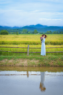 Beautiful Vietnamese Asian woman wearing a blue Ao Dai National Costume Dress with beautiful flowers stands relaxing beside yellow rice field with mountains in the background.