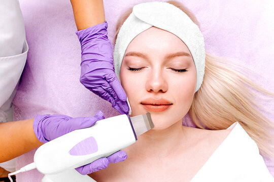 Young woman with a headband receives an ultrasonic peeling treatment on her face. Cosmetologist exfoliates the skin with an ultrasonic spatula in a beauty salon. Ultrasonic skin cleansing procedure.