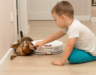 A little boy and a beautiful domestic leopard cat of the Bengal breed have fun playing in a home interior with a robot vacuum cleaner that cleans the apartment.