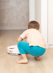 Cleaning concept. A small funny Caucasian boy, 4 years old, throws torn paper on the floor for cleaning by a white robot vacuum cleaner. Plays happily in the home interior.