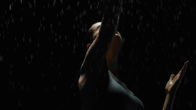 Silhouette elegant female dancing posing under rain at night. Seductive woman, gymnast dancer wearing black swimsuit, raindrops splashes, water drops roll down sexy body in shower, black background