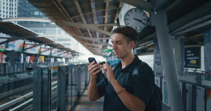 Man tourist with backpack using mobile phone while waiting for skytrain at metro station in big city. Man 30 tourist waiting skytrain at platform station of public transport. Urban traveling concept