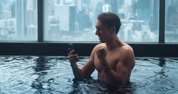 Happy Man 30s enjoys success read good news in smartphone while resting in luxury swimming pool on the roof of skyscraper on background of city. Mobile lottery win concept.