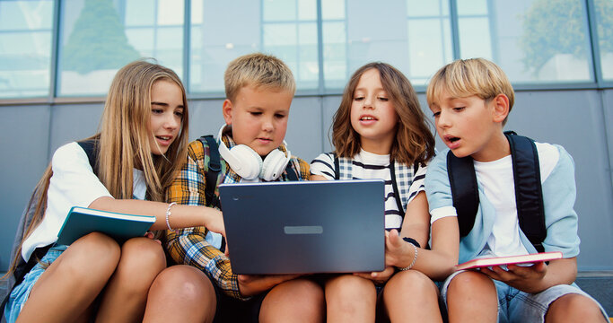 Back to school. Group of teenage school children using laptop sitting on stairs outside high school spending time together. Reading books, working with a laptop and communicating while sitting on the