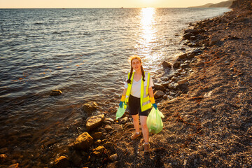 Top view of young Caucasian woman wearing vest and rubber holds plastic bag with garbage and bottle posing on pebble wild beach. Concept of Earth Day and save oceans