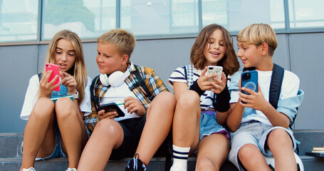 Cheerful stylish group of school children sitting on bench outdoors using and mobile phone communicating online. Friendship, technology concept. Playing game online