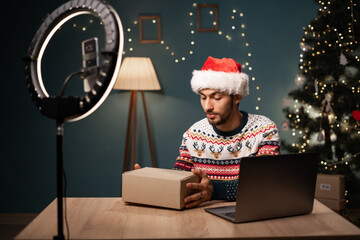 Happy arabic man social media channel blogger wear Santa hat recording unboxing parcel box, vlog on...