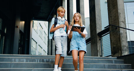Two teenage students boy and girl friends using digital tablet computer exchange lectures near school building summer day on stairs outside campus. Friendship. Back to school.