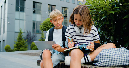 School students friends outdoors using digital tablet computer . Kids sitting on bench and play on laptop near school campus outdoors . Diverse kids doing a project together with tablet and notebook.