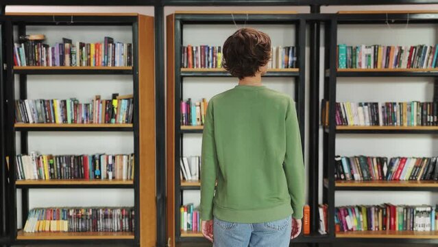 Back rear view young smart intelligent college student girl wearing green casual clothes studying visiting university library walk look for novel choosing book standing near shelf. Knowledge concept