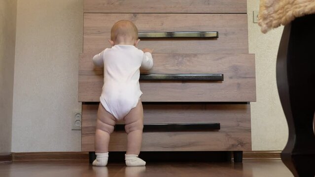 A little boy in a white bodysuit and socks stands near the chest of drawers and tries to open the drawer. A child starting to walk. Shot from the back. Overall plan. Slow motion. 4k footage