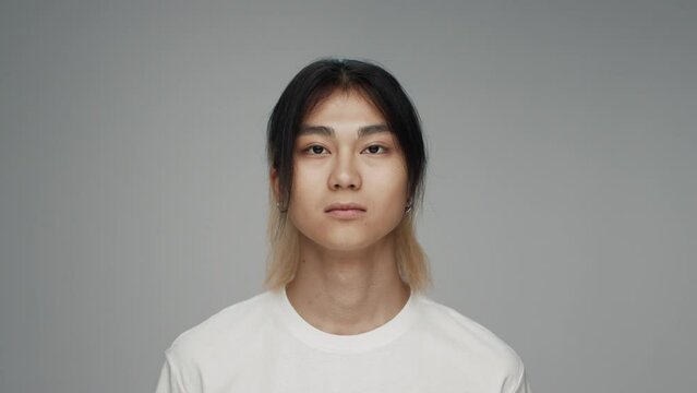 Portrait of Young Korean Man Looking at Camera in Colour Studio Shot. Adult Japanese Boy Isolated Alone on Grey Background Closeup. Asian 20s Person with Beautiful Eyes and Pensive Face Raising Head
