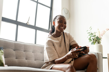 Excited young woman playing video games while sitting on sofa at home - obrazy, fototapety, plakaty