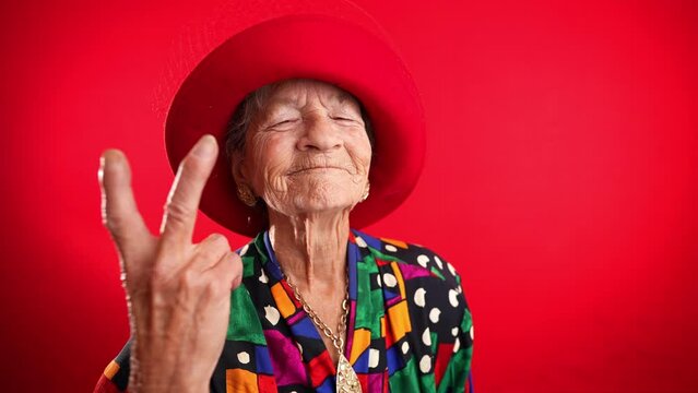 Portrait of funny, elderly mature woman, 80s, giving peace sign gesture with no teeth and red hat, isolated on purple background studio. Concept of youthful old lady with copy space