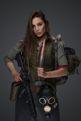 Middle Eastern-looking woman dressed in survivalist clothing posing with a rifle against a gray background, portraying strength and resilience