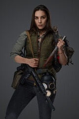 Naklejka premium Middle Eastern descent in post-apocalyptic survival gear, brandishing a dagger on a gray backdrop, showcasing her determination to survive