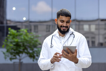 Smiling young male arab doctor standing outside on the street near the clinic and using a tablet