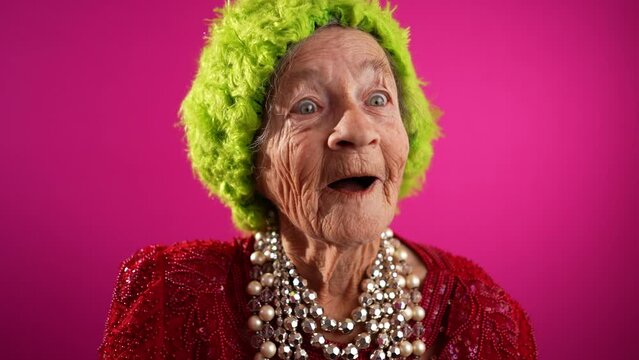Fisheye POV of funny mature elderly woman, 80s, having video chat with friends and family in front of computer, wearing green hat isolated on pink background.
