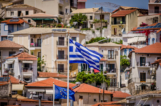 Greek Flag Waving in the Wind in Parga, Greece. Picturesque Coastal Town with Traditional Houses. Rooftops and Blue European Union Flag in Background.