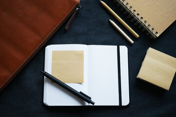 Open notepad with black pen next to leather brown laptop case, notepad and pens on dark background