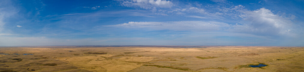 Fototapeta na wymiar Panoramic aerial view of a vast dry prairie landscape under a blue sky with white clouds. A dirt road cuts across the fields and a small pond reflects the blue sky. One very lonely tree sits in the ce