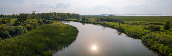 Panoramic view of a curving river that cuts through marshy land and is reflecting the sun. The sky has a thin overcast with scattered clouds.
