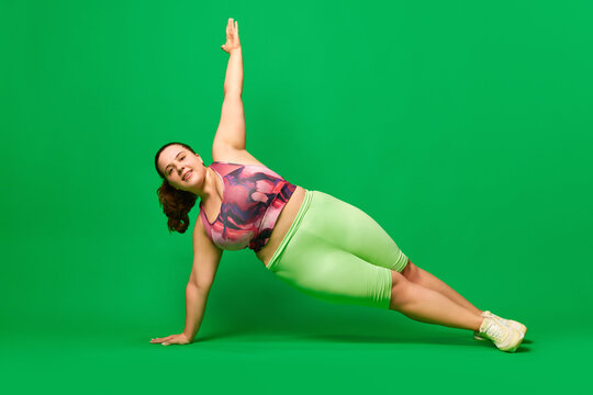 Positive, young overweigh woman training, standing in plank position against green studio background. Concept of sport, body-positivity, weight loss, body and health care. Copy space for ad