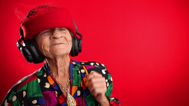 Funny happy old elderly woman with no teeth listening and dancing to music on smart phone in hand with headphones on red background