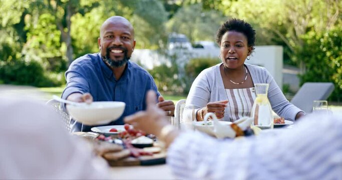 Family, lunch outdoor and eating in garden with drink, food and relax with conversation for thanksgiving. Black woman, African man and giving bowl for salad, meat and care at event, party or brunch