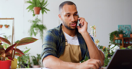 Businessman sitting among lots of green plants in plant shop, having talk on mobile phone and using laptop while taking an order from customer. Shop owner cheking online orders, e-mails, and typing