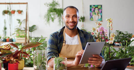 Portrait of african american man using digital tablet computer sitting wooden desk with botanic plants and looking into camera smiling. Male seller working at floral store checking order list on - obrazy, fototapety, plakaty