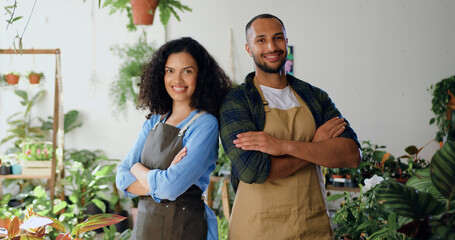Portrait of two young beautiful professional florist in aprons standing at own small flower shop and smiling to camera. African american couple posing in front of camera concept of family business and - obrazy, fototapety, plakaty