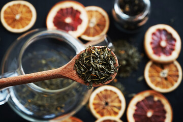 Loose leaf tea in a wooden spoon on the background of a glass teapot with tea and dry citrus slices