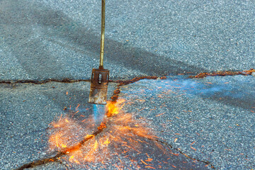 Asphalt patcher performs restoration work in cracks of road surface by burning dry grass before...