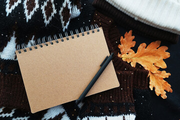 Notepad with black pen on a cozy sweater next to a hat and oak leaves