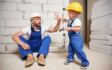 Male builder and little boy slapping hands giving high five in room with brick wall. Kid in safety...