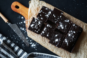 Chocolate brownie on a wooden board next to a knife and a towel on a dark background