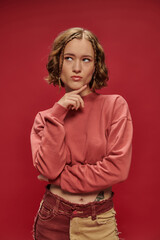 pensive stylish and young woman in patchwork pants and cropped long sleeve posing on red backdrop