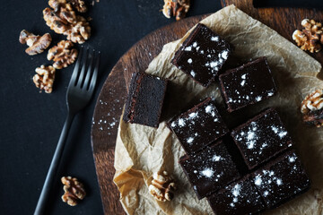 Chocolate brownie next to walnuts and a black fork on a wooden board on a dark background